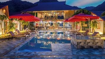 A pool area with lounge chairs and umbrellas, a building with a tiled roof, and a mountainous backdrop.