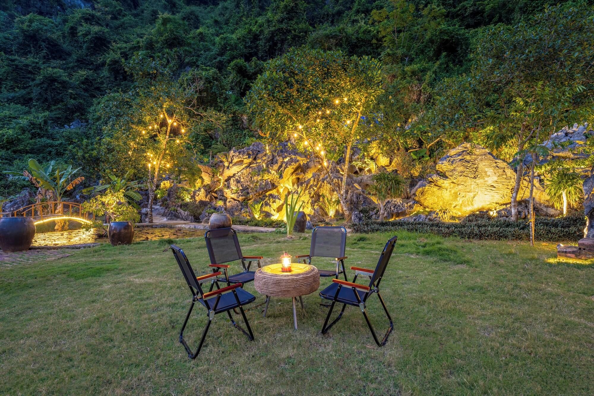An outdoor seating area with chairs and a table, surrounded by lush greenery and illuminated by string lights.