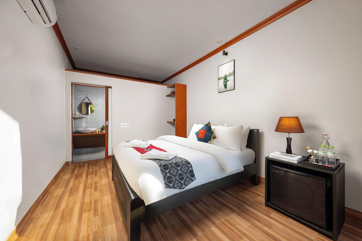 Una habitación de hotel moderna con piso de madera, una cama con ropa blanca, una mesita de noche con lámpara y un televisor pequeño.