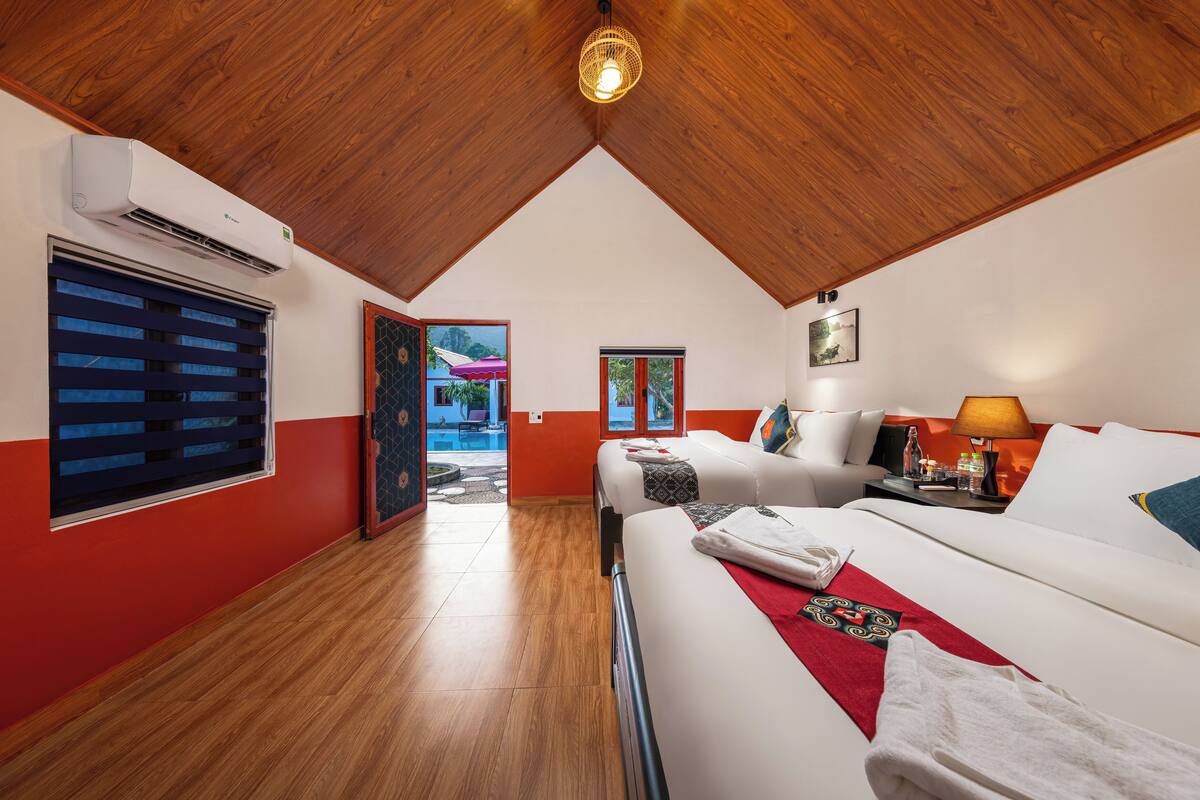 Une chambre d’hôtel avec un plafond en bois, un lit recouvert de draps blancs, un mur d’accent rouge et une fenêtre avec des stores.