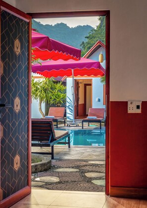 An outdoor pool area with lounge chairs and umbrellas, a view of a mountain, and a patterned wall. - Cat Ba Serena Homestay And Swimming Pool (Hai Phong Municipality)