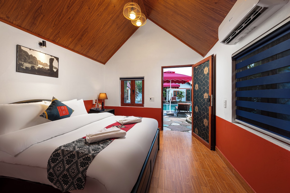 Een hotelkamer met een houten plafond, een bed met witte lakens, een nachtkastje met een lamp en uitzicht op een buitenzwembad.