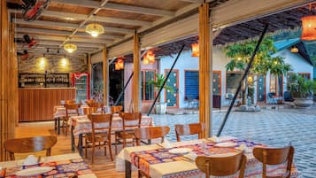 An outdoor restaurant with wooden tables and chairs, a bar area, and potted plants.