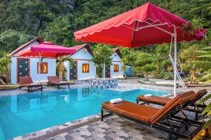 Zona de piscina con sillas de descanso y sombrillas, rodeada de edificios pequeños y abundante vegetación.