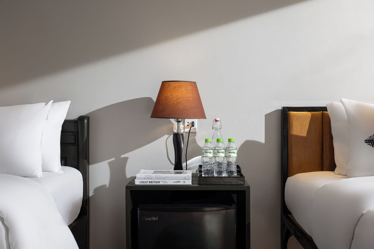 Habitación de hotel con dos camas blancas, una mesita de noche con lámpara, un refrigerador pequeño y una bandeja con agua embotellada y un libro.