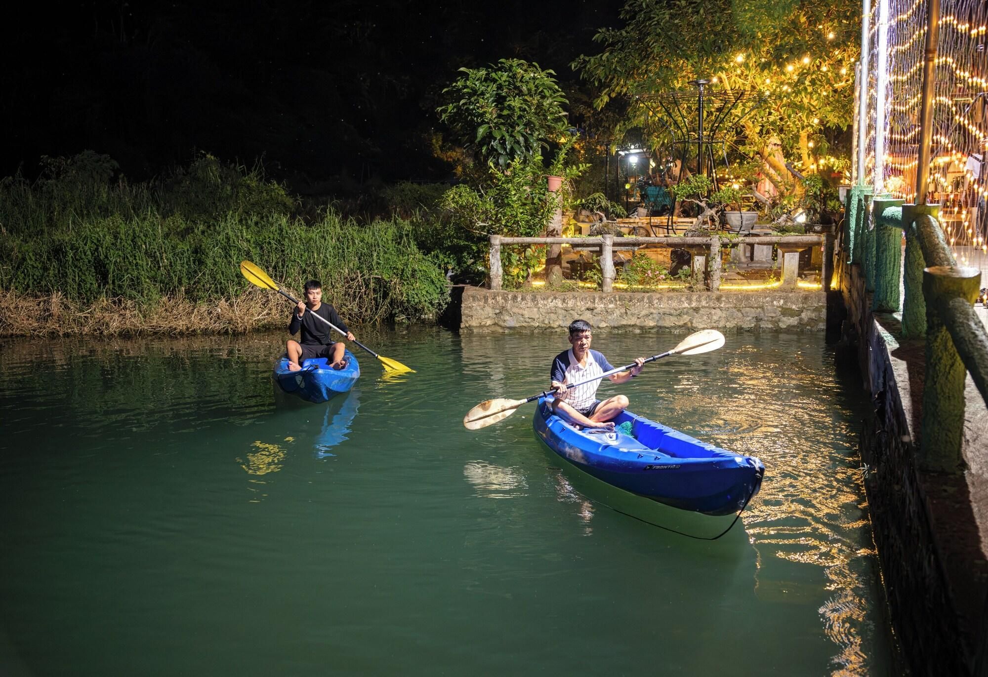 Two people kayaking on a calm river at night with a well-lit dock and greenery in the background.