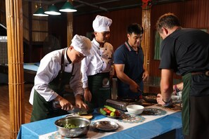Een groep koks bereidt eten in een keuken met bamboe kolommen en een blauwe tafelkleed.