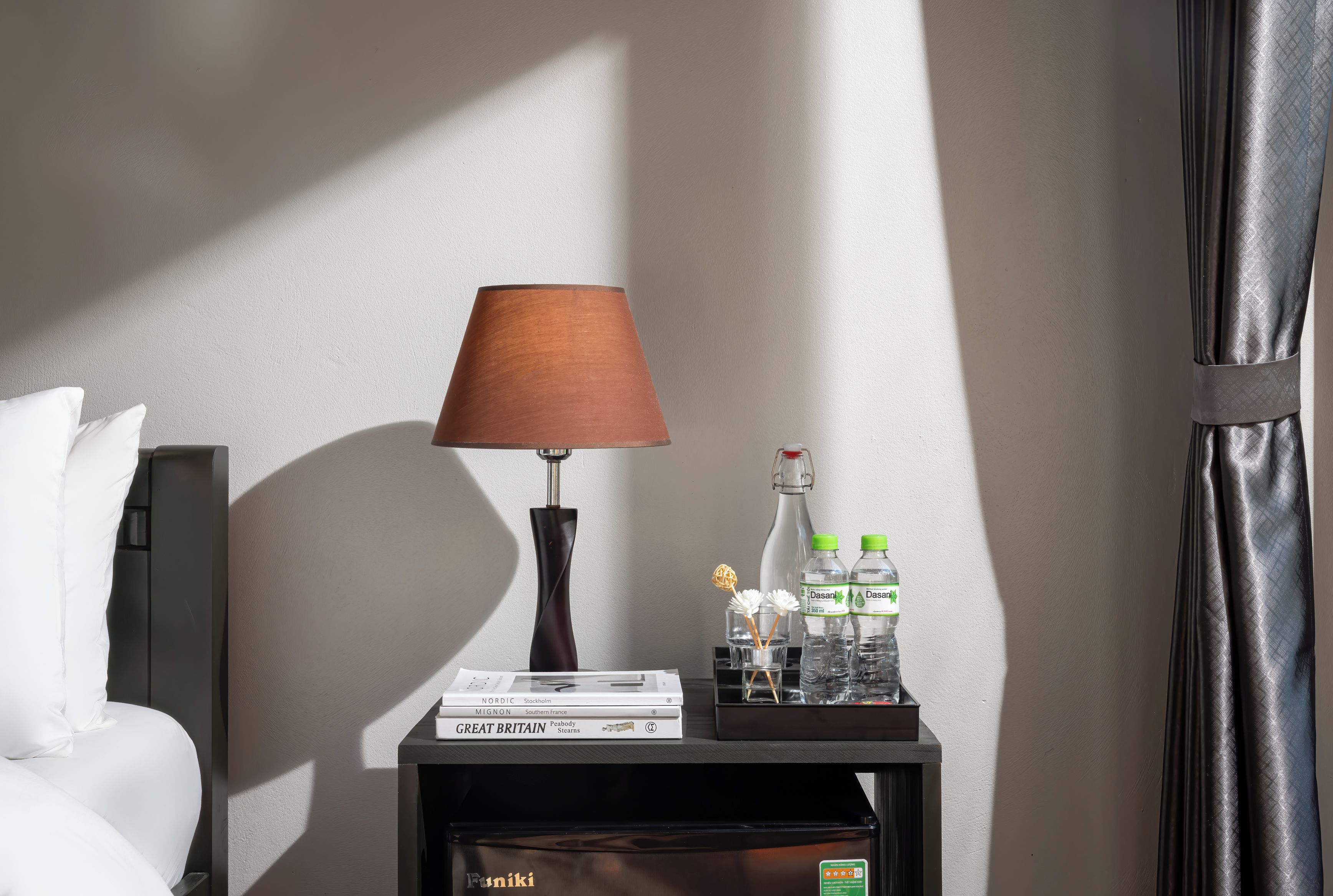 A neatly arranged hotel room with a bedside table featuring a lamp, a stack of books, and bottled water.