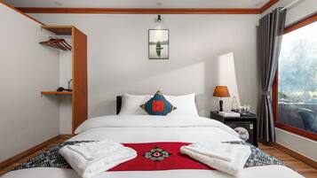 A hotel room with a neatly made bed, a bedside table with a lamp, a window with curtains, and a wall-mounted shelf with a hanging rack.