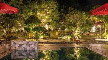 A nighttime outdoor area with a swimming pool, red umbrellas, and trees illuminated by string lights.