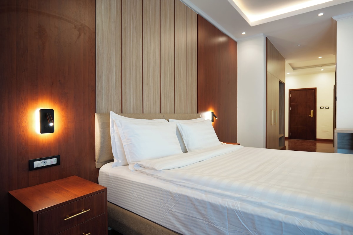 Cama bem arrumada com roupa de cama branca, mesa de cabeceira de madeira com gavetas e iluminação fixada na parede.