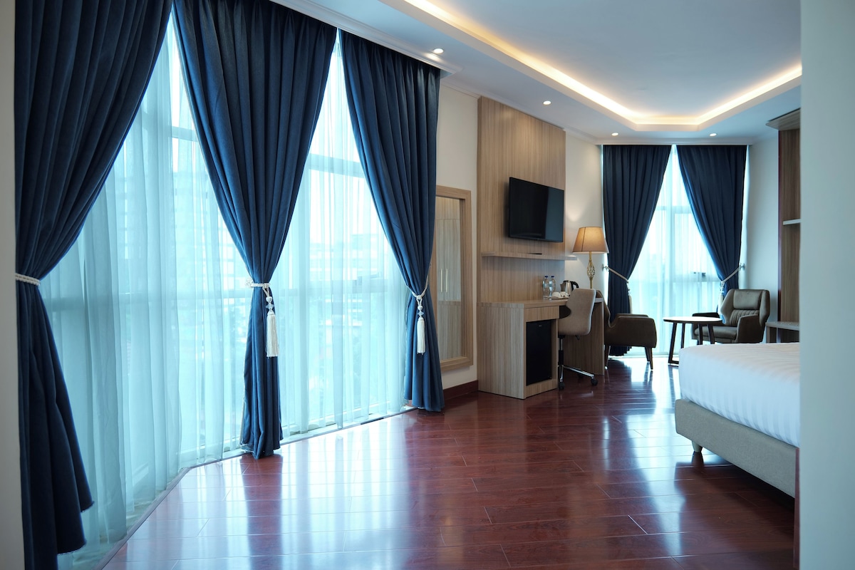 Quarto de hotel com uma cama grande, uma escrivaninha com cadeira, televisão na parede e janelas amplas com cortinas.