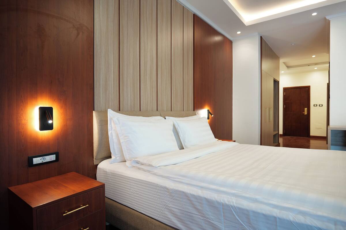 A neatly made bed with white linens, a wooden nightstand with drawers, and wall-mounted lighting.