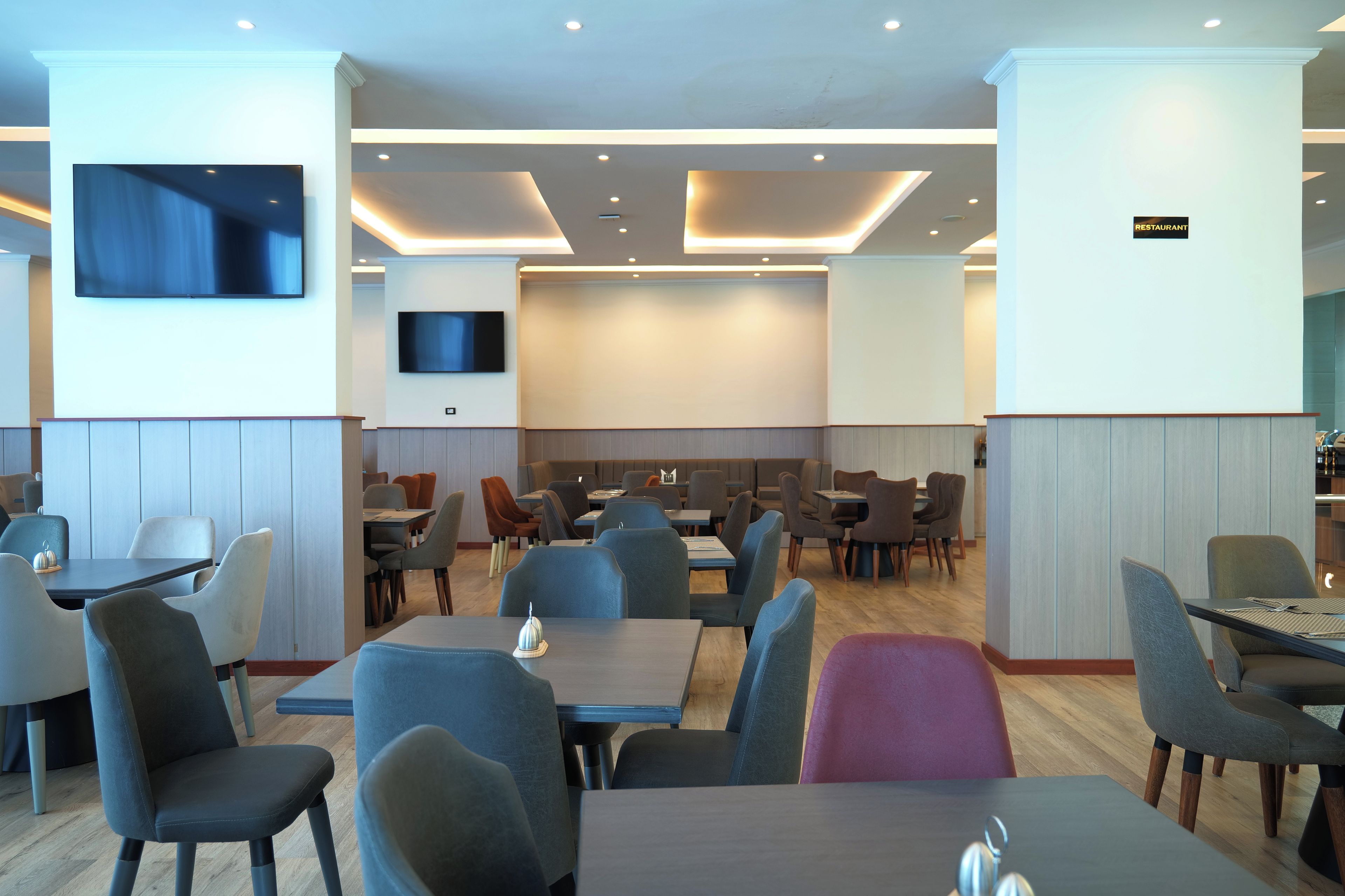 A modern restaurant interior with multiple dining tables and chairs, recessed ceiling lighting, and a flat-screen TV mounted on the wall.