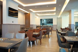 Un espace repas moderne avec plusieurs tables et chaises, un téléviseur à écran plat fixé au mur et un coin cuisine à l’arrière-plan.