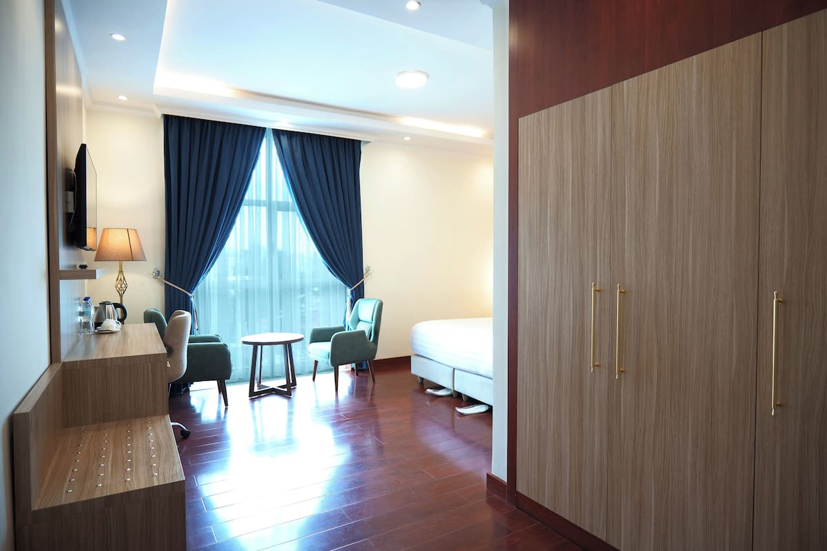 A hotel room with a wooden wardrobe, a bed, a desk with a lamp, two chairs, and a small table.