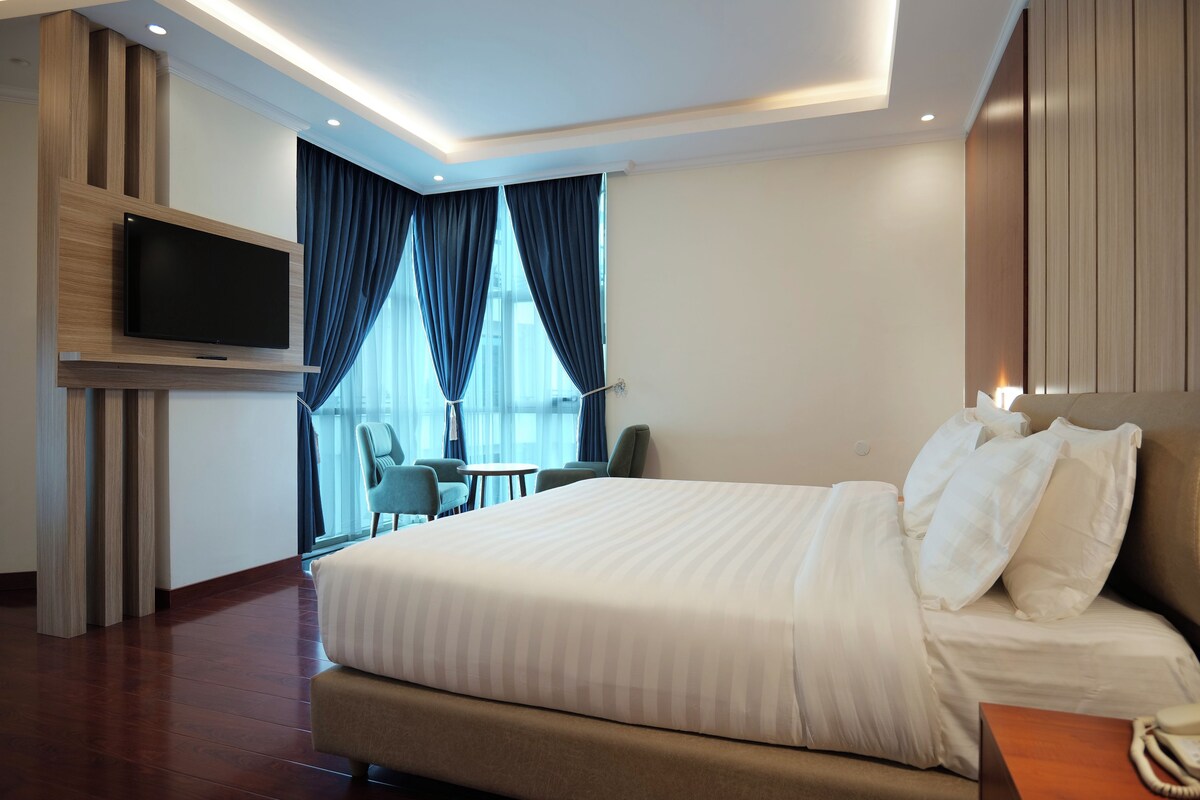 A modern hotel room with a large bed, a flat-screen TV mounted on the wall, a small table with chairs, and a view of the outdoors through large windows.