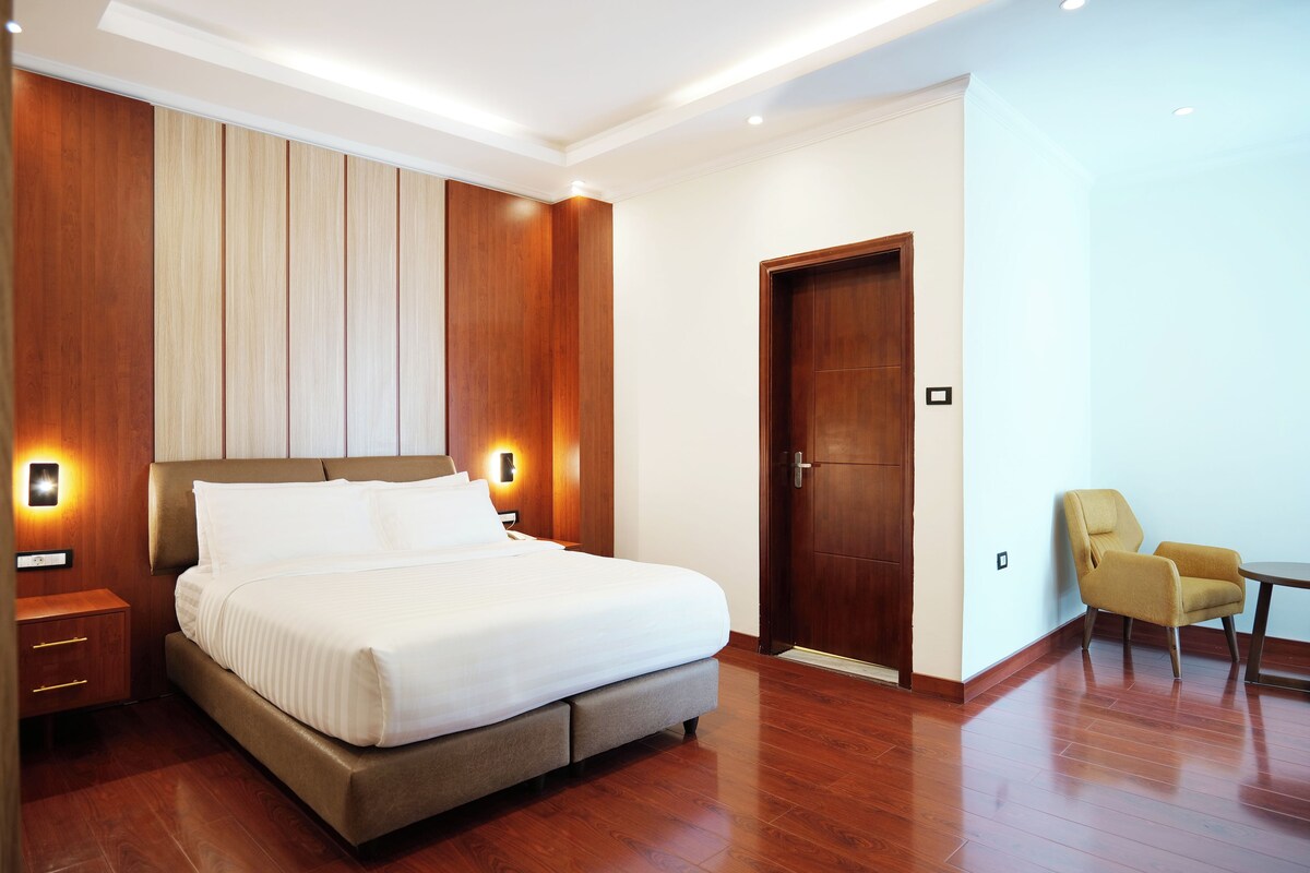 A modern hotel room with a large bed, wooden paneling, a wooden door, and a small table with a chair.