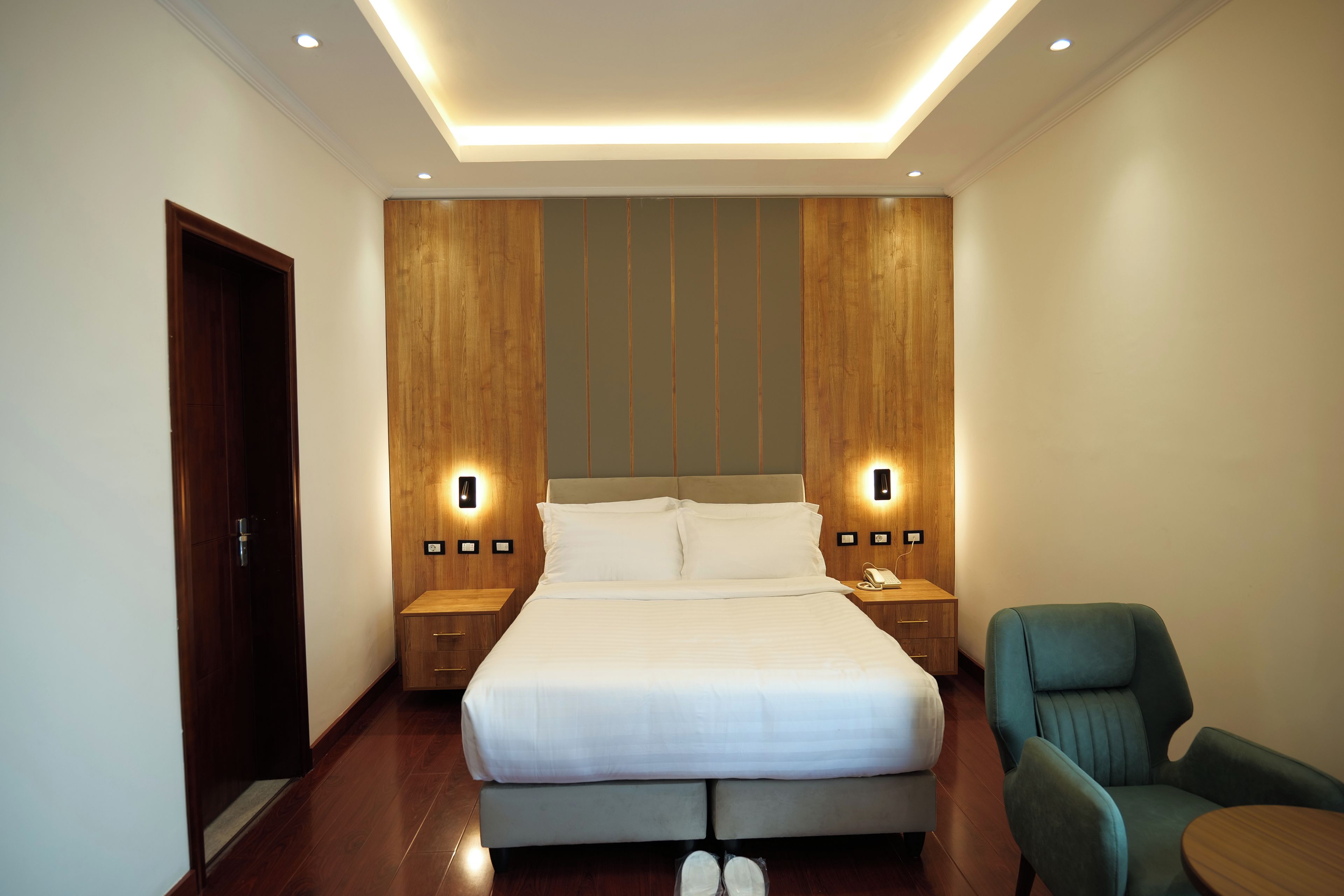 A hotel room with a large bed, two bedside tables with lamps, a wooden headboard, and a green armchair.