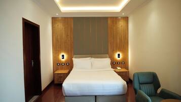 A hotel room with a large bed, two bedside tables with lamps, a wooden headboard, and a green armchair.