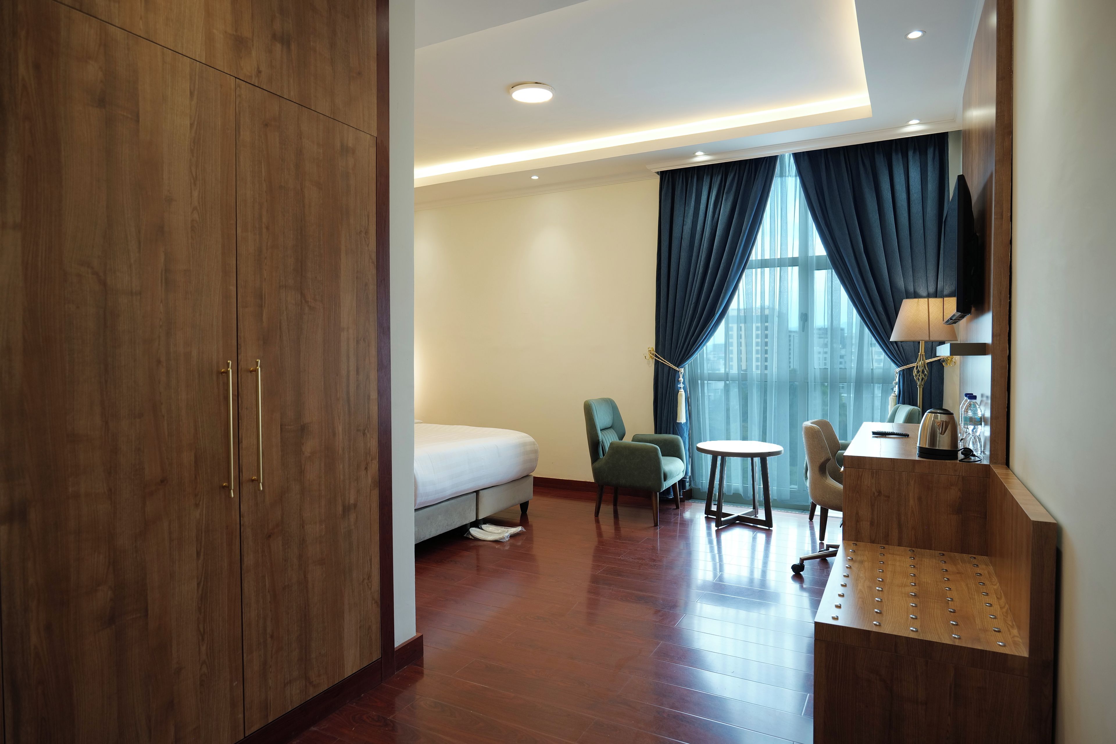 Quarto de hotel com cama, duas cadeiras, escrivaninha, luminária e uma janela grande com cortinas.