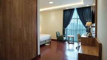 Quarto de hotel com cama, duas cadeiras, escrivaninha, luminária e uma janela grande com cortinas.