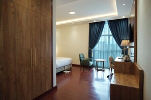 Quarto de hotel com cama, duas cadeiras, escrivaninha, luminária e uma janela grande com cortinas.