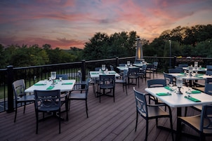 Un área de comedor al aire libre con mesas y sillas, rodeada por una terraza de madera y una barandilla.