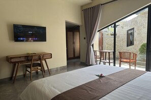 A hotel room with a bed, a desk with chairs, a TV mounted on the wall, and a view of the outside through a glass door. - Nawan Nusa Dua Bali By Zuzu (Kutuh)