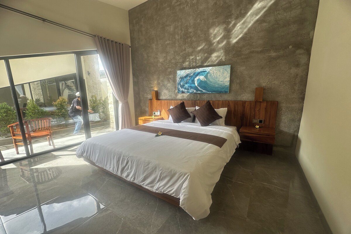 A modern hotel room with a large bed, a wooden headboard, a painting of a wave, and a balcony with a view.