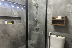 A modern bathroom with a glass-enclosed shower, grey marble tiles, a toilet, and a shelf with toiletries. - Nawan Nusa Dua Bali By Zuzu (Kutuh)