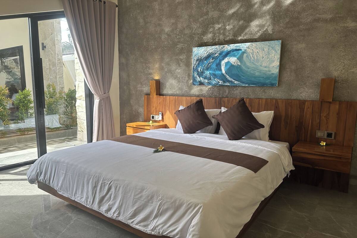 A large bed with a wooden headboard, a nightstand, and a wall-mounted painting of a wave.