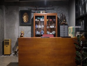 A room with a wooden reception desk, a Buddha statue, a cabinet with decorative items, and a fan. - Another High Away Co.,ltd (Bang Kruai)