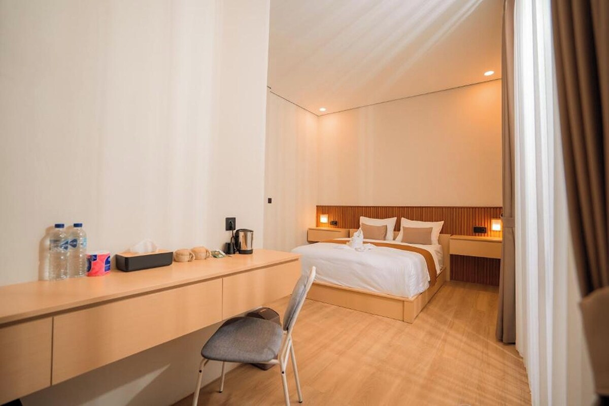 A modern hotel room with a wooden desk, a bed with white linens, and a small bedside table with a lamp.