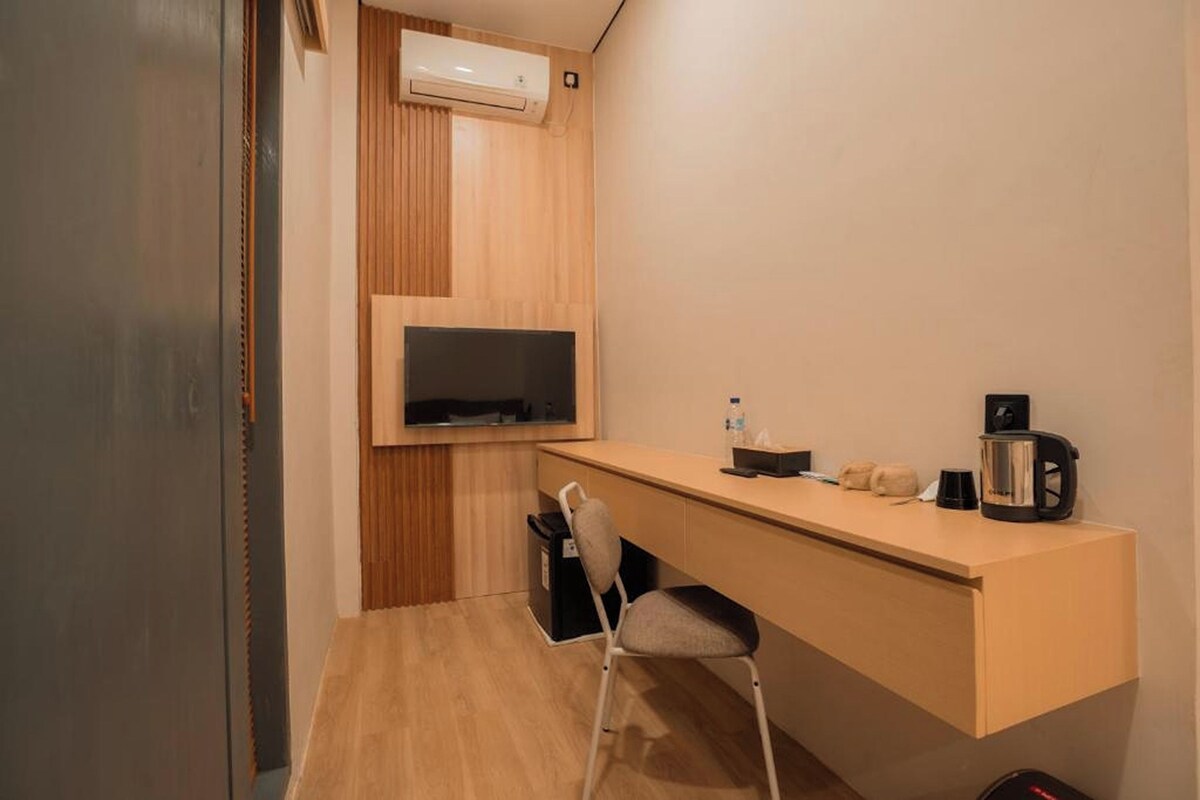 Ein kleines Hotelzimmer mit Schreibtisch aus Holz, einem Sessel, einem Fernseher und einer Kaffeemaschine.