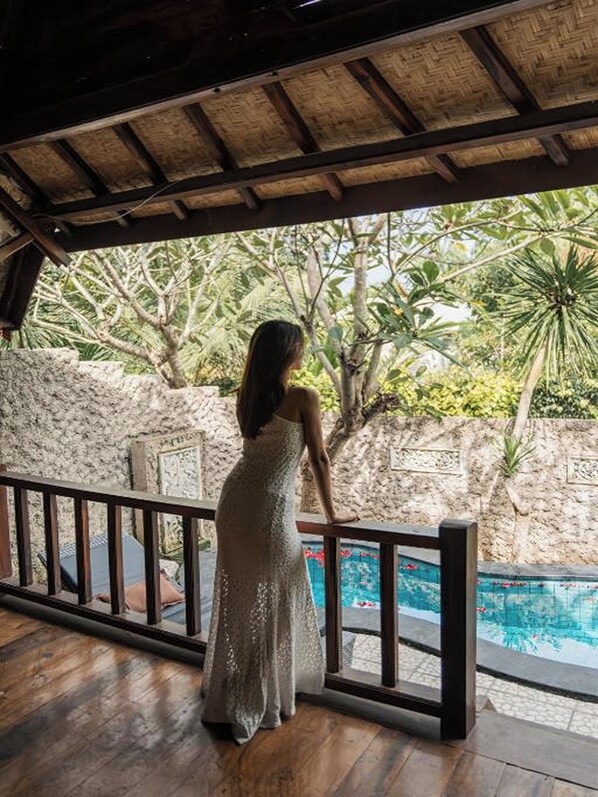 Eine Frau in einem weißen Kleid steht an einem Pool, im Hintergrund eine Steinmauer und tropische Pflanzen.