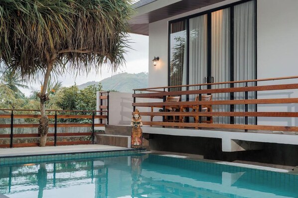 Un espace piscine moderne avec une terrasse en bois, une statue et une vue sur les collines.