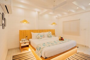 A modern bedroom with a wooden headboard, a large bed with a floral bedspread, two hanging lamps, a nightstand, and a window with blinds. - Elivaas House Of Neptune (Morjim)