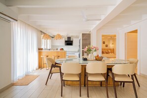 A modern dining area with a glass table, six chairs, and a kitchen in the background. - Elivaas House Of Neptune (Morjim)