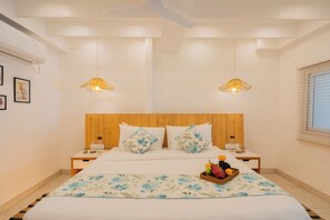 Une chambre avec une tête de lit en bois, deux tables de chevet, un lit avec une literie à motifs floraux, deux lampes fixées au mur et un panier de fruits posé sur le lit.