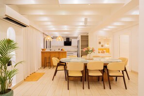A modern dining area with a round glass table, beige chairs, and a kitchen in the background. - Elivaas House Of Neptune (Morjim)