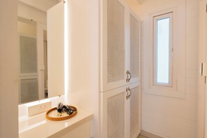 A bathroom with white cabinets, a mirror, a window, and a basket with hairdryers on the counter. - Elivaas House Of Neptune (Morjim)