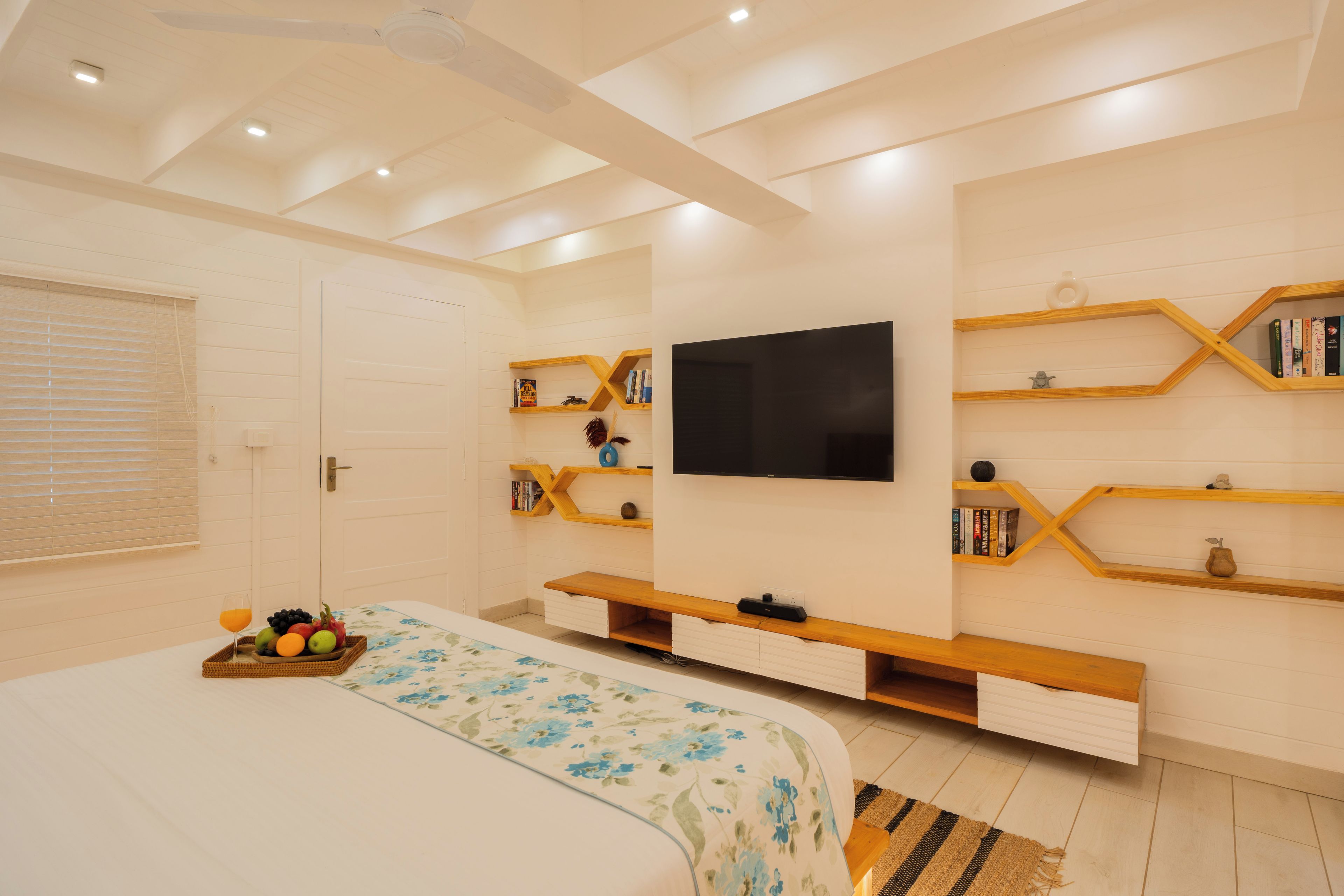 Un dormitorio con una cama, un televisor montado en la pared, estanterías de madera y una cesta de frutas.