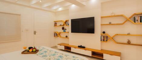 Un dormitorio con una cama, un televisor montado en la pared, estanterías de madera y una cesta de frutas.