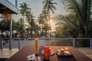 A table with a drink, a mug, and a tray of snacks, set against a backdrop of palm trees and a sunset. - Elivaas House Of Neptune (Morjim)