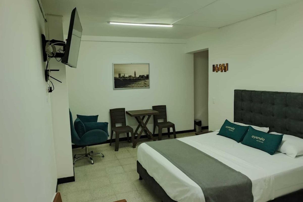 Habitación de hotel con una cama, un escritorio, dos sillas, un televisor y un cuadro en la pared.