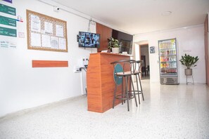 Zona de recepción con escritorio de madera, dos taburetes altos, un televisor montado en la pared, un tablón de anuncios con documentos y una máquina expendedora.