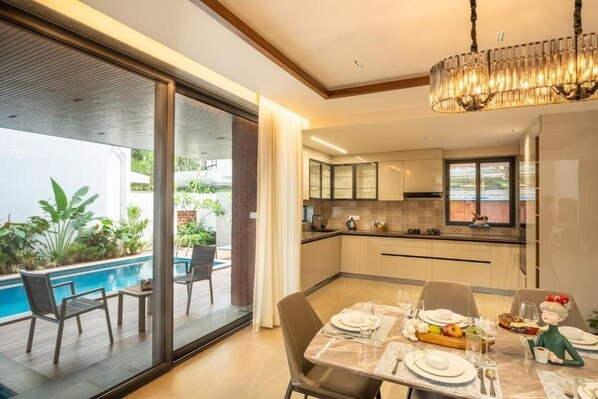 A modern kitchen with a dining area, a pool, and a view of greenery. - Elivaas Cloud 7-luxury Villa In Goa (Vagator)