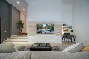 A modern living room with a large TV mounted on a wooden wall, a seating area with a sofa and ottoman, and a small dining table with chairs.