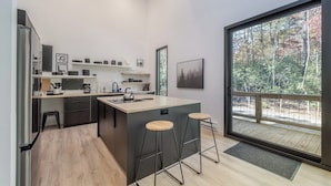 A modern kitchen with a central island, bar stools, and a large window offering a view of trees.